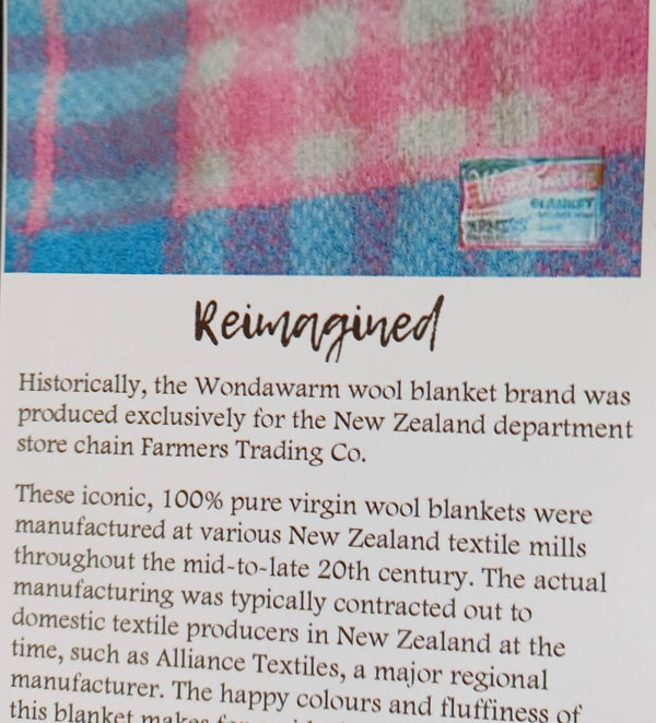 A printed card or booklet page with a small inset photo of a pink and blue patterned wool blanket above a block of text headed 'Reimagined.' The page is photographed against a dark fabric background.